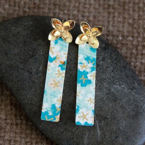 Peut inclure: Une paire de boucles d'oreilles rectangulaires en métal doré et turquoise avec un motif floral. La partie turquoise des boucles d'oreilles présente un motif floral.