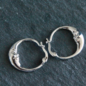 May include: A pair of silver hoop earrings featuring a crescent moon design. Each earring has a small clear stone set in the centre of the moon.