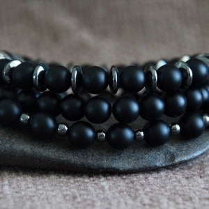 May include: Three black beaded bracelets with silver accents. The bracelets are stacked on top of each other and are resting on a grey stone.