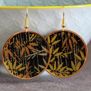 May include: A pair of gold hoop earrings with a black and gold fabric design. The fabric has a pattern of leaves and branches.