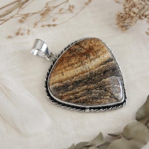 May include: A triangular pendant featuring a polished brown and tan landscape jasper stone with dark brown horizontal banding. The stone is set in a silver-toned metal frame with a twisted rope border and a smooth, polished silver-toned bail at the top.