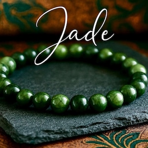 Puede incluir: Un brazalete de jade con cuentas redondas y pulidas en varios tonos de verde. El brazalete se exhibe sobre una superficie oscura y texturizada, con la palabra "Jade" en cursiva arriba. El fondo presenta una tela estampada.