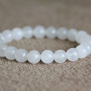 May include: White gemstone bracelet with round beads. The bracelet is made of white stones and is on a brown fabric background.