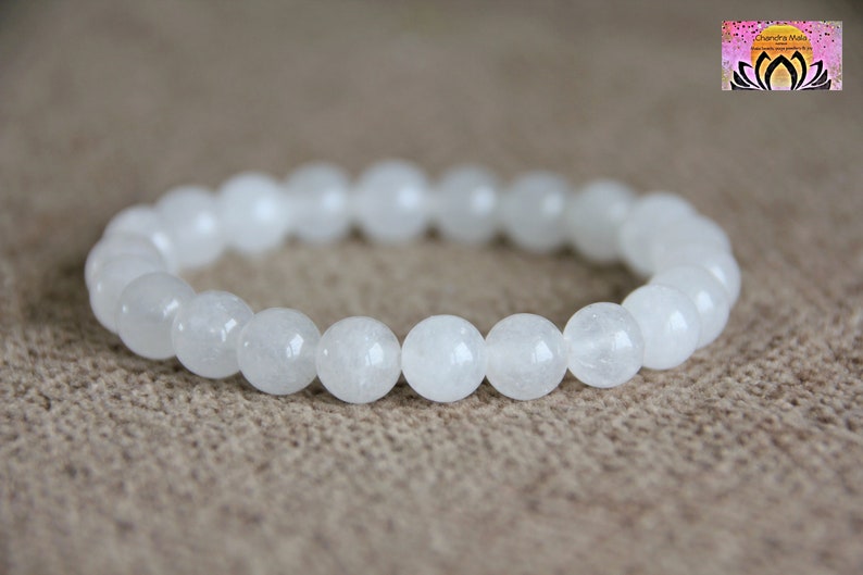 May include: White gemstone bracelet with round beads. The bracelet is made of white stones and is on a brown fabric background.