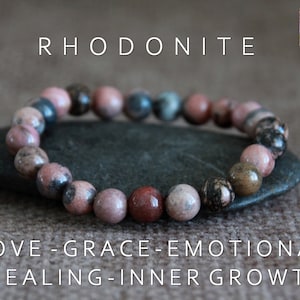 May include: Rhodonite beaded bracelet with pink, brown, and gray stones. The bracelet is displayed on a dark gray stone, with text that reads "LOVE - GRACE - EMOTIONAL HEALING - INNER GROWTH".