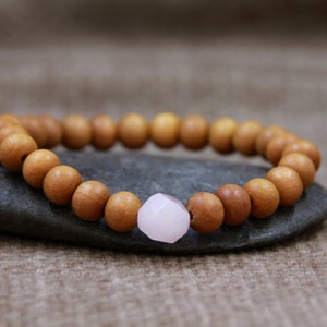 May include: A wooden beaded bracelet with a pink rose quartz bead. The bracelet is on a grey rock.