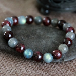 May include: A bracelet made with alternating dark red and gray beads. The bracelet is on a gray stone.