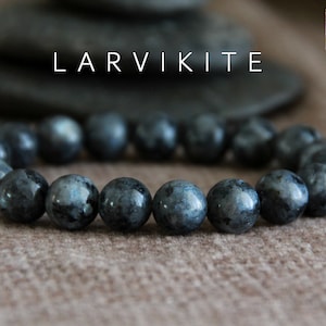 Puede incluir: Un brazalete de piedra Larvikite negra con cuentas redondas. El brazalete está sobre una superficie marrón texturizada. El texto "LARVIKITE" está en letras blancas sobre el brazalete.