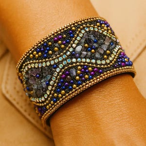 May include: A wide cuff bracelet featuring a black base adorned with intricate patterns of small gold, blue, and purple beads. The design includes embedded grey stone fragments and rows of iridescent crystals, framed by gold-toned metal edges.
