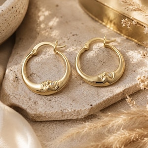 May include: A pair of gold hoop earrings featuring a raised celestial face design on the bottom curve of each hoop. Each earring includes a small, clear, round gemstone embedded within the cheek area of the face motif and a hinged post closure.
