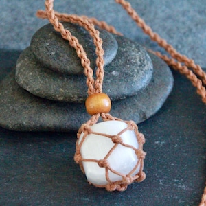 May include: A white crystal pendant wrapped in brown macrame cord, hanging from a brown cord necklace. The pendant is resting on a stack of grey stones.