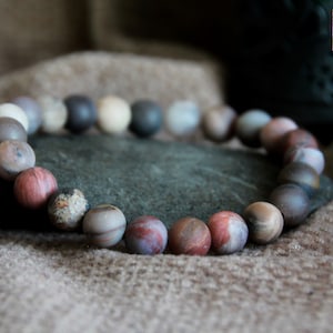 Puede incluir: Un brazalete hecho de cuentas de piedras preciosas marrones, grises y rojas. Las cuentas son redondas y tienen un acabado liso y pulido.