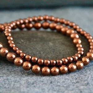 May include: Three copper beaded bracelets stacked on top of each other. The beads are round and have a shiny finish.