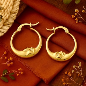 Könnte beinhalten: Ein Paar goldfarbene Creolen. Jeder Ohrring hat ein Mond-Design mit einem Gesicht und einem kleinen, klaren Stein. Die Ohrringe werden auf einem rostfarbenen Stoff mit floralen Akzenten präsentiert.