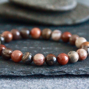May include: A beaded bracelet featuring a variety of earth-toned beads. The beads are primarily shades of brown, red, and cream, with variations in texture and pattern. The bracelet is displayed on a dark gray surface.