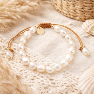 May include: A pearl bracelet with a tan cord and gold accents. The bracelet features a series of white, irregularly shaped pearls strung together. A small gold disc and pearl-shaped beads adorn the bracelet.