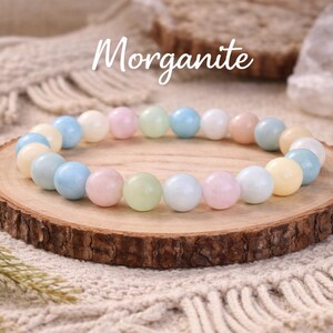May include: A beaded bracelet featuring smooth, round stones in pastel shades of pink, blue, yellow, green, and white. The word Morganite is written in white script above the bracelet, which is arranged in a circle on a round wooden slice.