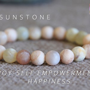 Può includere: Un braccialetto in pietra di sole con perline rotonde di colore beige chiaro, marrone chiaro e bianco. Il braccialetto ha le parole "JOY - SELF EMPOWERMENT - HAPPINESS" scritte sotto.