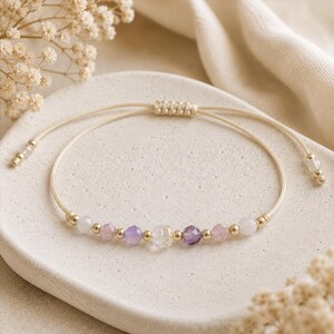May include: A delicate adjustable bracelet featuring a beige cord with a sliding knot closure. The centre is adorned with a sequence of small gold-toned beads interspersed with faceted translucent beads in shades of white, pale pink, and purple.