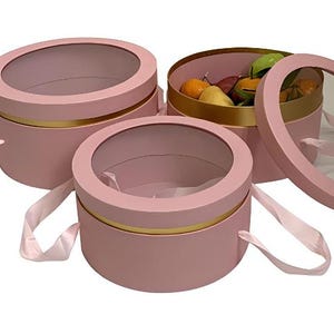 May include: Three pink round gift boxes with clear circular windows and gold trim. Each box has a light pink ribbon handle. One box is open, revealing fruit inside. The boxes are designed for gifting or storage.