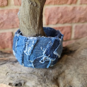 May include: A close-up of a wooden branch with a denim fabric planter around it. The planter is made of blue denim fabric with frayed edges.