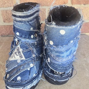 May include: A pair of denim boots with a patchwork design. The boots are made from blue denim with white polka dots and feature a black interior lining. The boots have a distressed, frayed edge design and the OshKosh B'gosh logo is visible.
