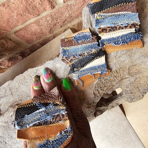 May include: Handmade patchwork leather and denim accessories. The pieces feature a mix of brown, black, and blue denim, with leopard print accents. The edges are stitched with contrasting thread, creating a unique, textured look.