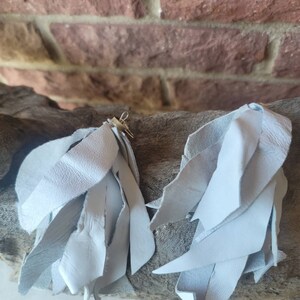 May include: A pair of light gray leather tassel earrings. The tassels are made of thin strips of leather that are frayed at the ends.