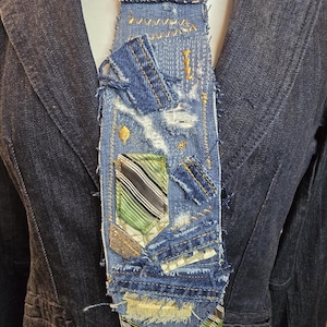 May include: A denim tie with a patchwork design, featuring various shades of blue denim, distressed details, and decorative stitching. The tie includes a pocket with green and gray stripes. The tie is worn with a dark blue blazer.