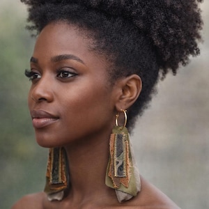 May include: Gold hoop earrings with olive green and brown fabric pendants. The earrings have a layered design with stitched details. The woman in the image has dark skin and is wearing a black top.