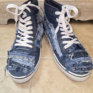 May include: A pair of high-top denim sneakers with white laces. The blue denim shoes have a distressed, patchwork design with frayed edges and metal stud accents. The soles are white with a black stripe.