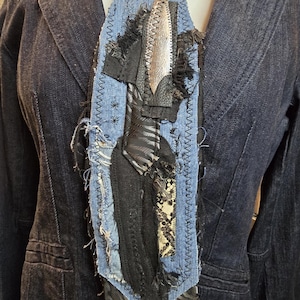 May include: A unique denim necktie with a patchwork design. The tie features a central black fabric element with a silver accent, surrounded by frayed denim and decorative stitching. The tie is worn over a dark denim jacket.