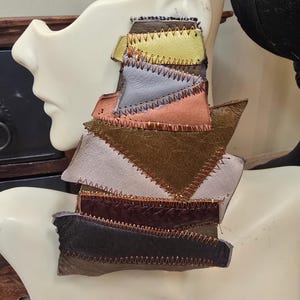 May include: A large, asymmetrical earring composed of layered leather patches in various colors, including olive green, gray, and peach. The patches are stitched together with contrasting gold thread, creating a unique, geometric design. The earring is displayed on a mannequin head.