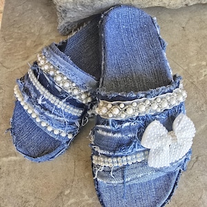 May include: A pair of denim slides with white pearl embellishments and a white pearl bow. The slides are distressed and have a frayed edge.