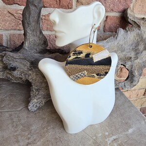 May include: A pair of large, round earrings made of leather with a patchwork design in shades of brown, black, white, and gold. The earrings are hanging from silver hoops.