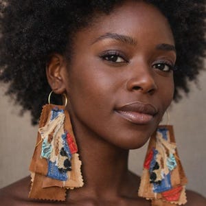 May include: A woman with dark skin and curly black hair wears large, layered earrings. The earrings are made of brown suede and feature patches of blue, red, and cream-colored fabric with decorative stitching. The earrings hang from gold hoops.