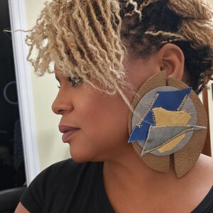 May include: A large, circular statement earring featuring a textured tan leather base with a split design. The center is layered with irregular, stitched patches of blue, grey, and yellow leather, creating a multi-dimensional, geometric appearance.