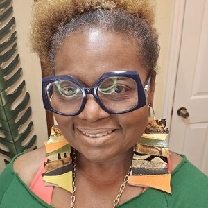 May include: A woman with short, curly, blonde hair is wearing large, dark blue, square-shaped glasses and a green shirt. She is wearing long, colorful, fabric earrings with gold accents.