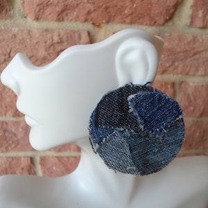 May include: A large, round earring made from patches of denim fabric in various shades of blue. The earring is attached to a white mannequin head, showcasing the design and texture of the repurposed denim material. The earring has a unique, handcrafted appearance.