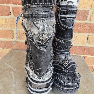 May include: A pair of black denim boots with decorative stitching and embellishments. The boots have a distressed look with patches of lighter fabric and silver-toned metal accents. The soles are gray.
