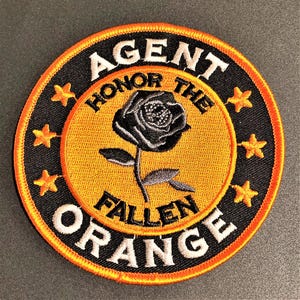 May include: A black and orange embroidered patch with the text "AGENT ORANGE HONOR THE FALLEN" and a black rose with a gray stem. Five white stars surround the circle.