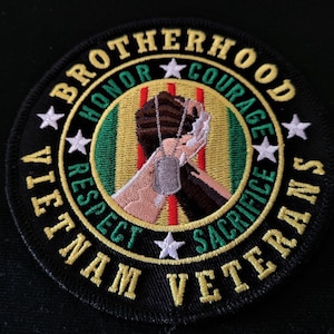 May include: A black and yellow embroidered patch with the words "Vietnam Veterans Brotherhood" around the edge. Inside the circle is a depiction of two hands clasped together, one white and one black, holding a dog tag. The words "Honor, Courage, Respect, Sacrifice" are also embroidered around the hands.