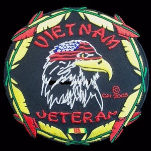 May include: A black and yellow embroidered patch with a white eagle wearing a red, white, and blue bandana. The eagle's head is facing right. The words "Vietnam Veteran" are embroidered in red and white around the eagle. The patch is surrounded by a green and yellow wreath.