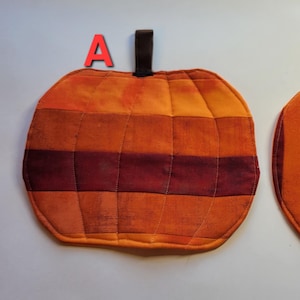 May include: Two pumpkin-shaped quilted pot holders in shades of orange and burgundy. Each has a brown loop for hanging. One is striped horizontally, the other vertically. The letter "A" and "B" are above each pot holder.