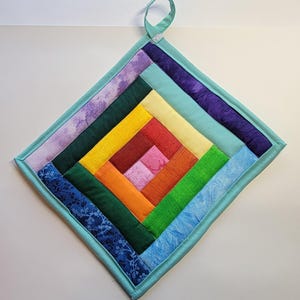 May include: A square, quilted potholder with a rainbow log cabin design. The center is pink, surrounded by red, orange, yellow, green, blue, and purple fabric strips. A light blue border and hanging loop complete the design.