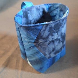 May include: A cylindrical fabric storage basket featuring a quilted construction with a blue tie-dye pattern. The exterior displays a geometric patchwork design in various shades of navy and royal blue, complete with a small fabric loop handle.