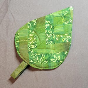 May include: A quilted leaf-shaped pot holder in shades of green. The fabric features a mix of solid and patterned sections with a leaf design. A small fabric loop extends from the base. The pot holder is designed for kitchen use.