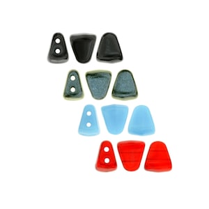 May include: A collection of colorful glass beads in various shapes and colors. The beads include black, green, light blue, and red, with some featuring two holes for stringing. The beads are arranged in rows against a white background.