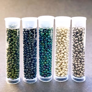 May include: Five clear plastic tubes filled with small, cylindrical beads in shades of green, blue-green, beige, and gray. Each tube has a white cap. The beads appear to be for hair extensions or crafting.