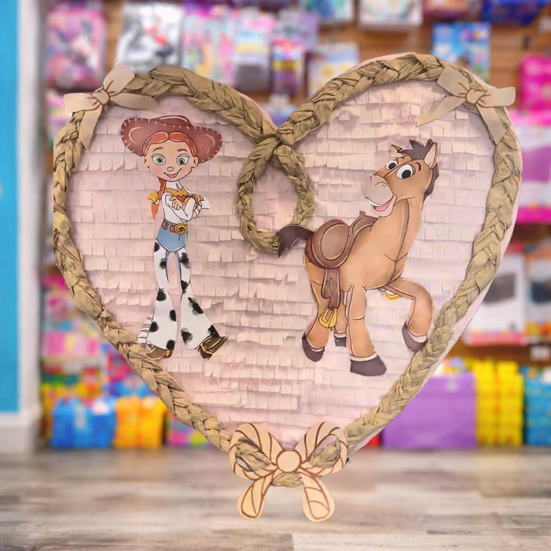 May include: Heart-shaped pi&ntilde;ata featuring cartoon characters Jessie and Bullseye from Toy Story. The pi&ntilde;ata has a light pink background with a braided rope border and a decorative bow at the bottom. The characters are in full color.